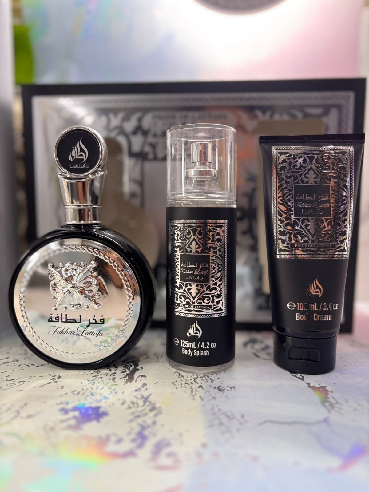 Perfumería oriental de estética oscura del catálogo Kikluxe Colombia 2026: set Lattafa Fakhar con frasco negro y medallón plateado, body splash 125ml y body cream 100ml — el universo oud de Emiratos que convive con el Ombre Nomade Louis Vuitton en el catálogo, envíos a 13 ciudades